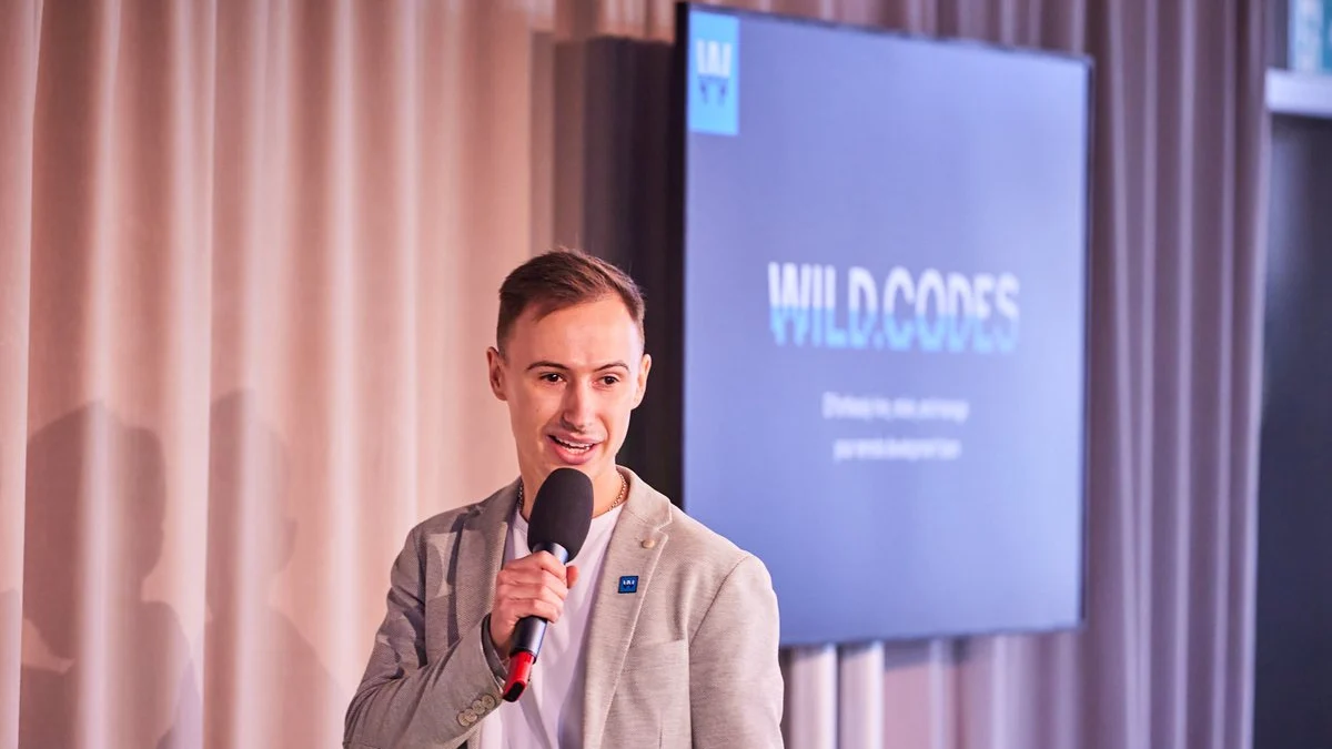 Serge Lavrinchuk, co-founder of Wild.Codes, speaking on stage with a microphone in front of a screen showing the Wild.Codes logo.