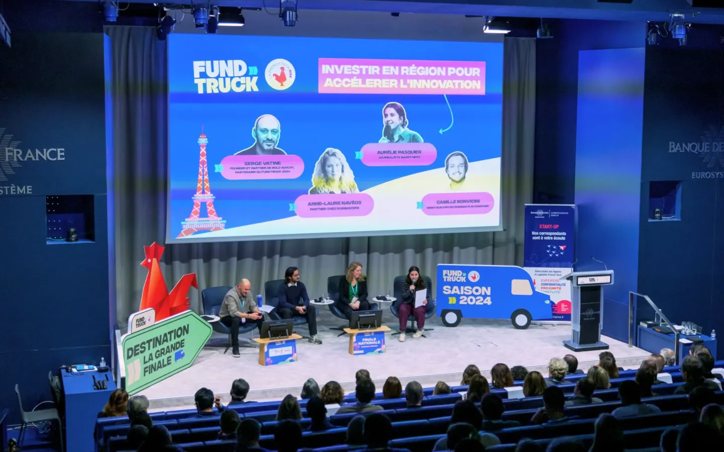 FundTruck 2024 startup pitch event in a conference hall. Panelists discuss regional investment and innovation, with a large screen displaying speakers' names and topics. Audience attentively listening.